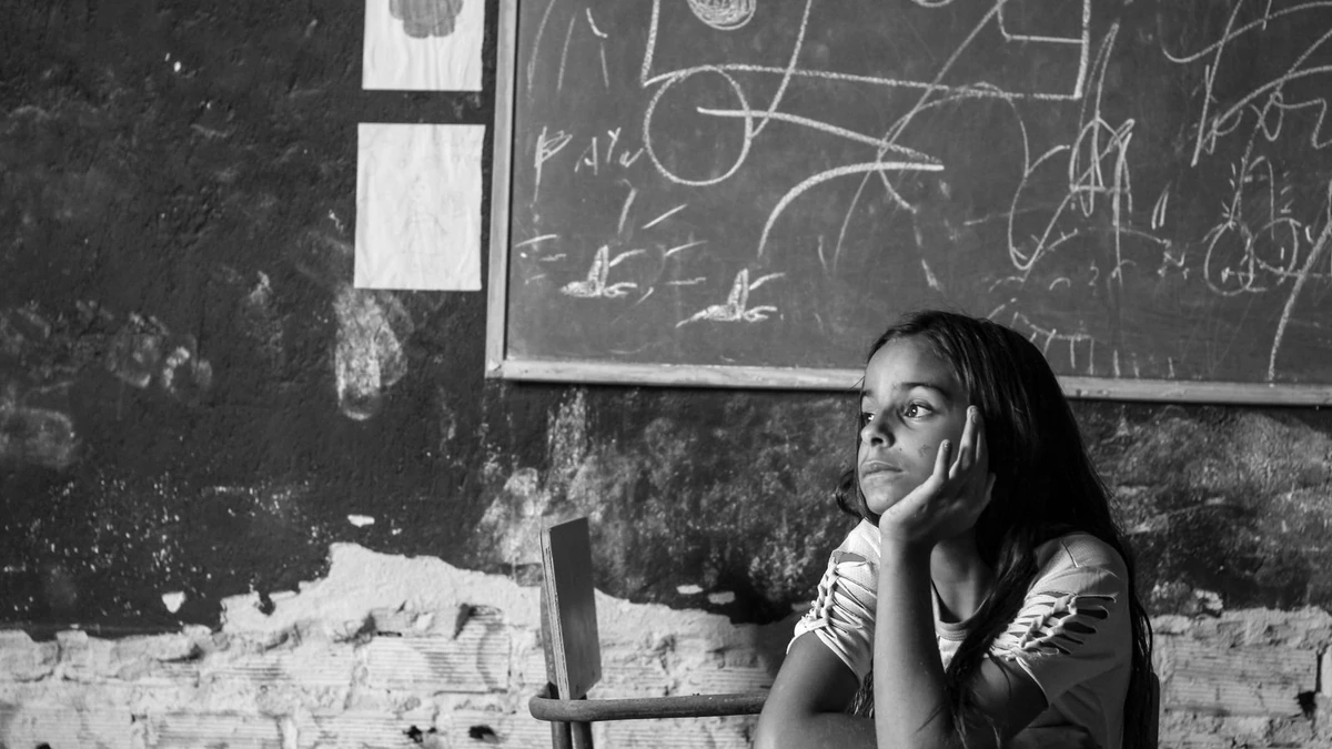 Menina brasileira pensativa sentada em sala de aula com quadros de giz ao fundo, tema do estudo sobre escolas de Salvador afetadas por tiroteios em dias letivos